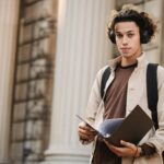 Young learner in wireless headphones in backpack standing with documents near entrance of university building