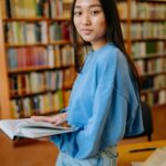 Asian woman reading a book in a library, smiling while studying surrounded by shelves.